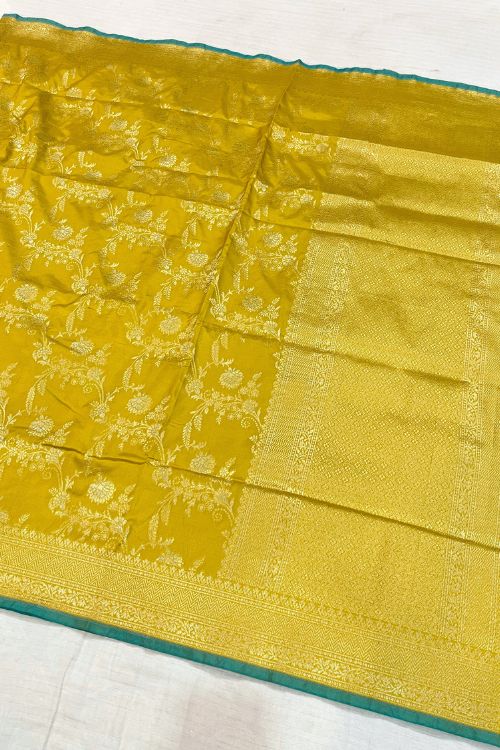 Handwoven Mustard Yellow Katan Silk Saree with intricate floral design, contrast green selvedge border, and detailed aachal. Paired with an unstitched lilac blouse with matching sleeve borders.