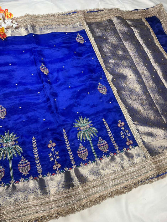 This royal blue hand-embroidered pure tissue silk saree reflects timeless Banarasi artistry and festive grandeur. Crafted in shimmering pure Banarasi tissue silk, the saree features delicate hand-embroidered kalash motifs, palm trees, floral vines, and ti