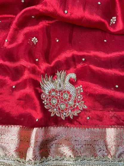 "Red 100% pure Banarasi tissue silk saree featuring a beautifully embroidered white swan motif with floral detailing in silver and red. Adorned with scattered buti work, a richly woven zari pallu, and a gota patti lace border. Ideal for weddings, receptions, and festive occasions. Comes with an unstitched brocade blouse."