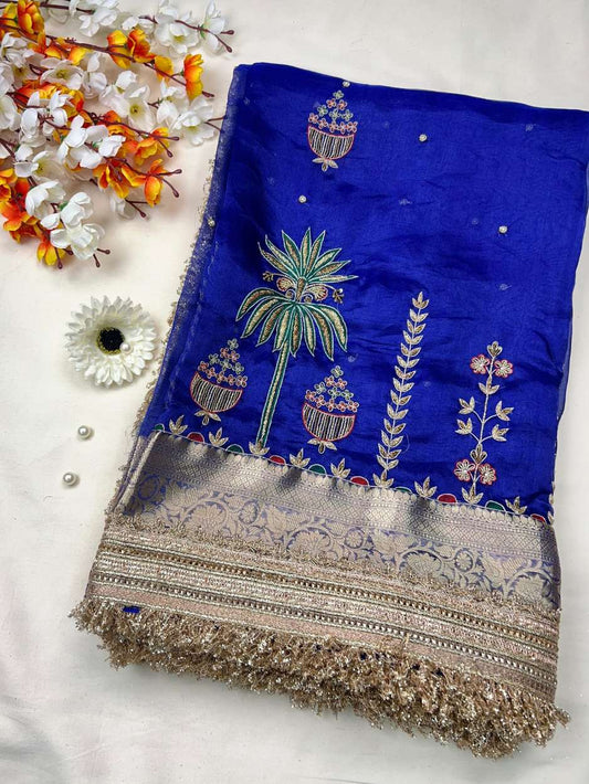 This royal blue hand-embroidered pure tissue silk saree reflects timeless Banarasi artistry and festive grandeur. Crafted in shimmering pure Banarasi tissue silk, the saree features delicate hand-embroidered kalash motifs, palm trees, floral vines, and ti