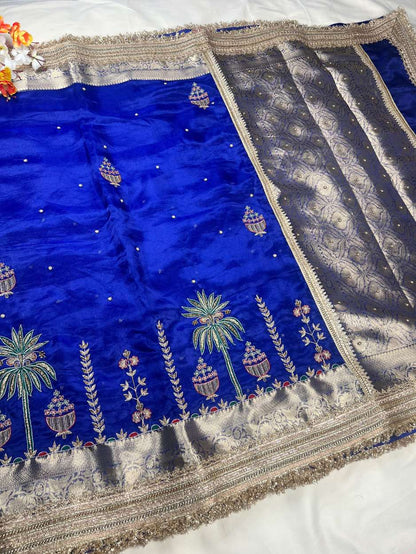This royal blue hand-embroidered pure tissue silk saree reflects timeless Banarasi artistry and festive grandeur. Crafted in shimmering pure Banarasi tissue silk, the saree features delicate hand-embroidered kalash motifs, palm trees, floral vines, and ti
