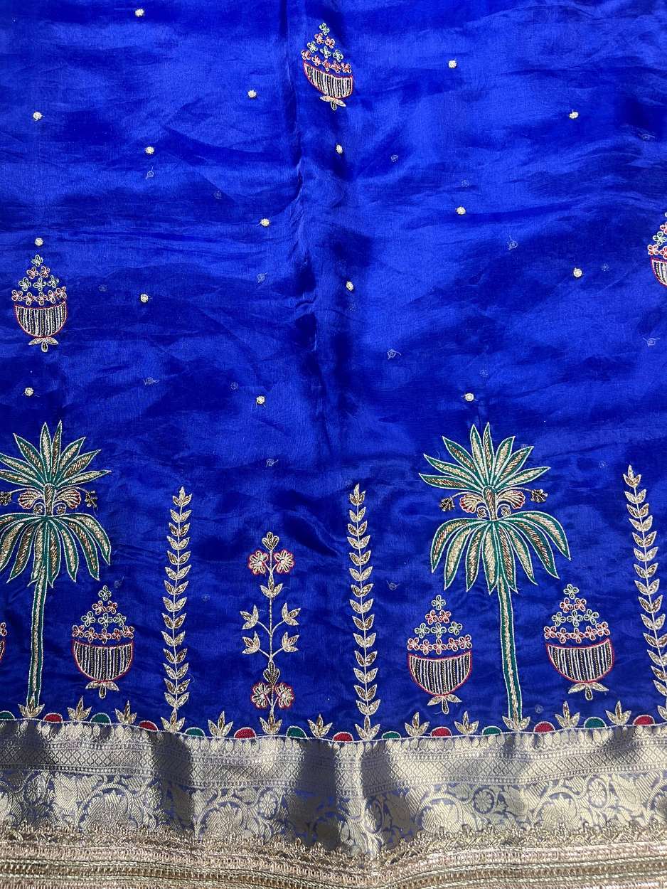 This royal blue hand-embroidered pure tissue silk saree reflects timeless Banarasi artistry and festive grandeur. Crafted in shimmering pure Banarasi tissue silk, the saree features delicate hand-embroidered kalash motifs, palm trees, floral vines, and ti