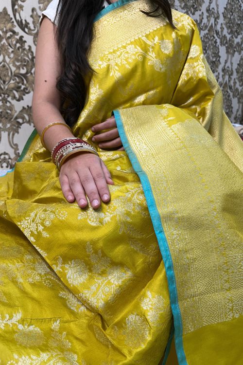 Handwoven Mustard Yellow Katan Silk Saree with intricate floral design, contrast green selvedge border, and detailed aachal. Paired with an unstitched lilac blouse with matching sleeve borders.